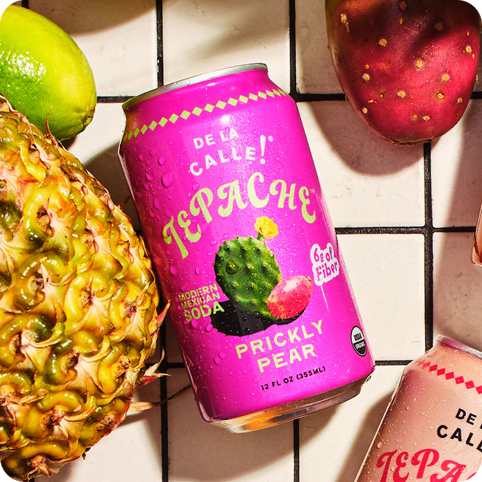 Pink can of De La Calle Tepache Prickly Pear soda with a pineapple and strawberry on a tiled surface.