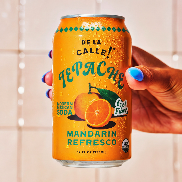 Orange can of De la Calle Tepache Mandarin Refresco held by a hand with blue nail polish against a neutral background.