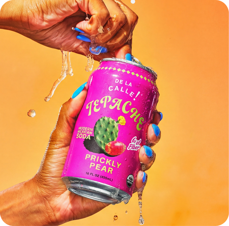 Person holding a can of Tepache Prickly Pear soda with a colorful label against an orange background
