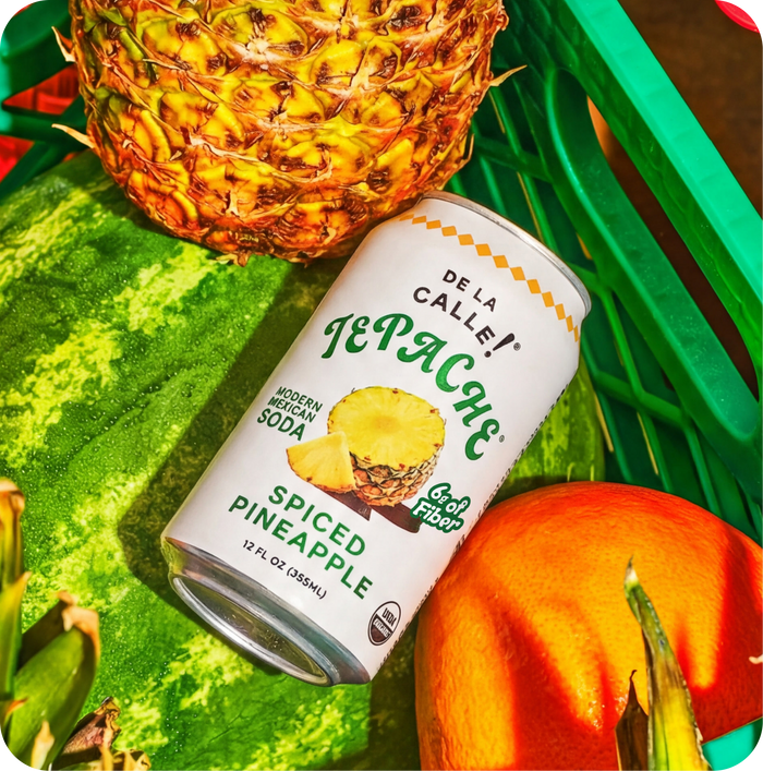 Can of De La Calle Tepache Spiced Pineapple Soda with a pineapple, watermelon, and pumpkin on a green surface.