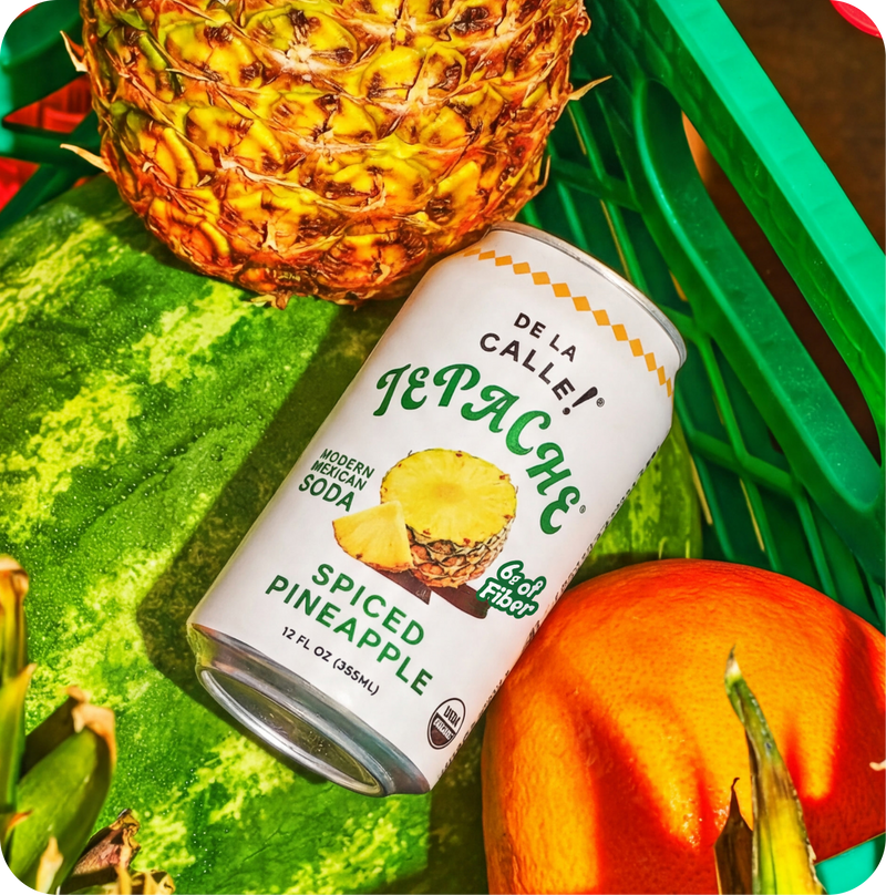 Can of De La Calle Tepache Spiced Pineapple Soda with a pineapple, watermelon, and pumpkin on a green surface.