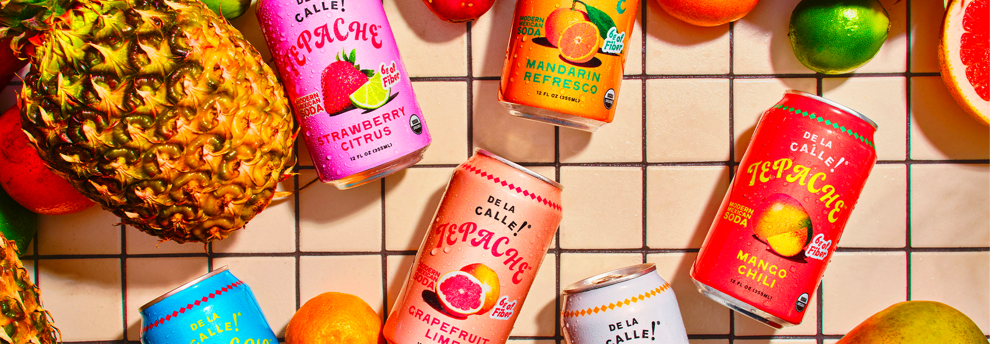 Assorted cans of de la calle tepache on a tiled surface with fruits around.