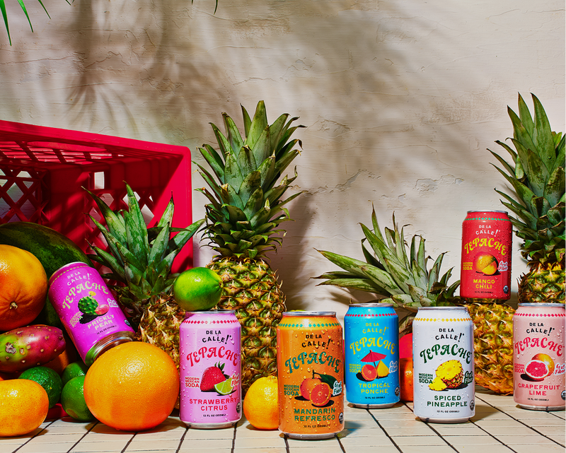 A group of de la calle fruit-flavored canned drinks arranged on a tabletop with pineapples, oranges, limes, and a red crate in a styled studio setup.