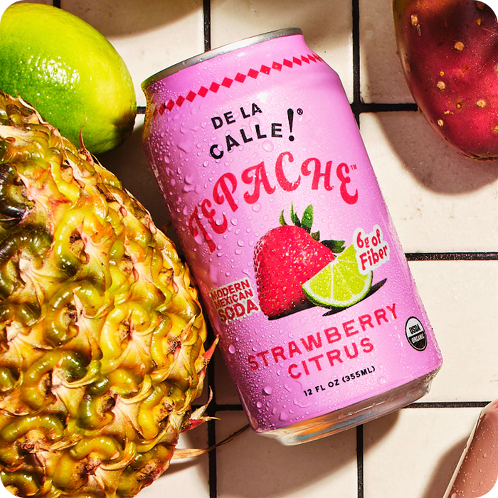 Pink can of Depache Strawberry Citrus soda with a pineapple, lime, and strawberry on a checkered surface.