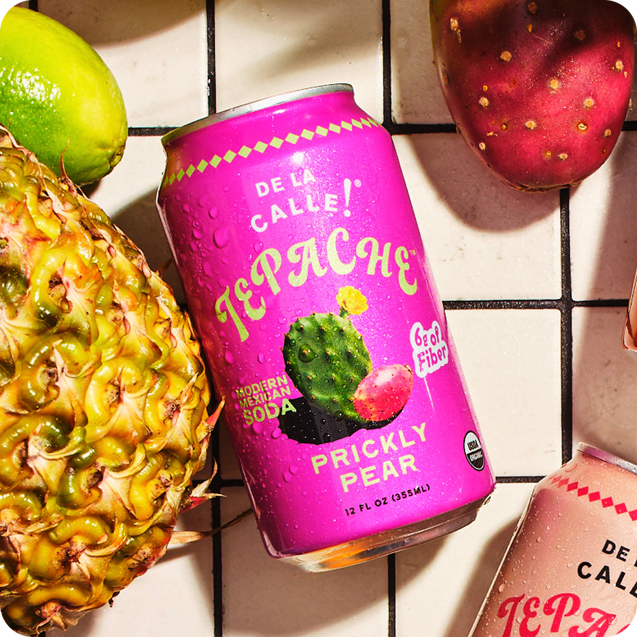 Pink can of De La Calle Tepache Prickly Pear soda with a pineapple and strawberry on a tiled surface.