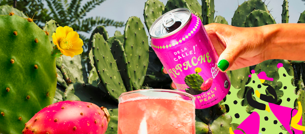 A hand with green nail polish holds a pink can of Dela Calle prickly pear Soda against a background of cacti and a glass of pink drink.
