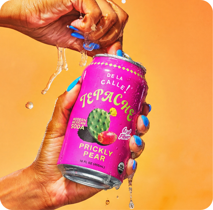 Person holding a can of Tepache Prickly Pear soda with a colorful label against an orange background